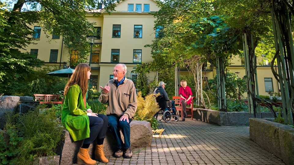 En kvinna och en äldre man sitter framför en byggnad som inramas av grönska. 