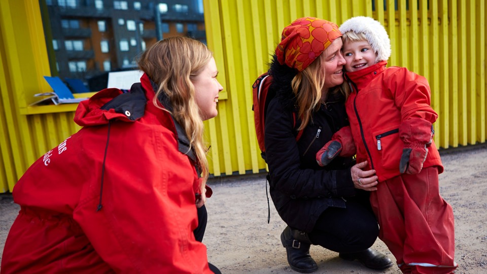 Ett barn i röd overall står lutad mot förälder och tittar mot pedagog som sitter på huk.