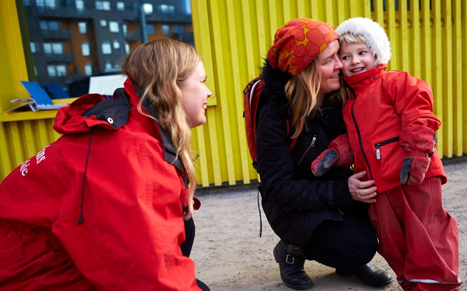 Ett barn i röd overall står lutad mot förälder och tittar mot pedagog som sitter på huk.
